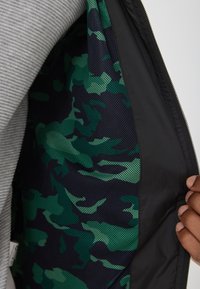 Sort jakke med et grønt og marineblåt camouflage-meshfor, med lynlås foran og et struktureret gråt stof i kanten.