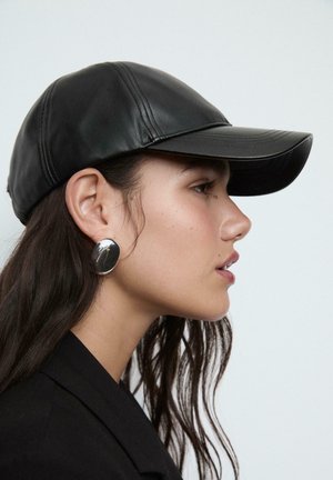 Cappello in pelle nera con visiera curva, caratterizzato da dettagli di cucitura; indossato con un grande orecchino circolare in argento su uno sfondo neutro.
