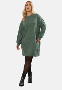 Abito maglione verde a maniche lunghe con vestibilità rilassata; presenta un collo rotondo e tasche laterali. Indossato con stivaletti neri alla caviglia.