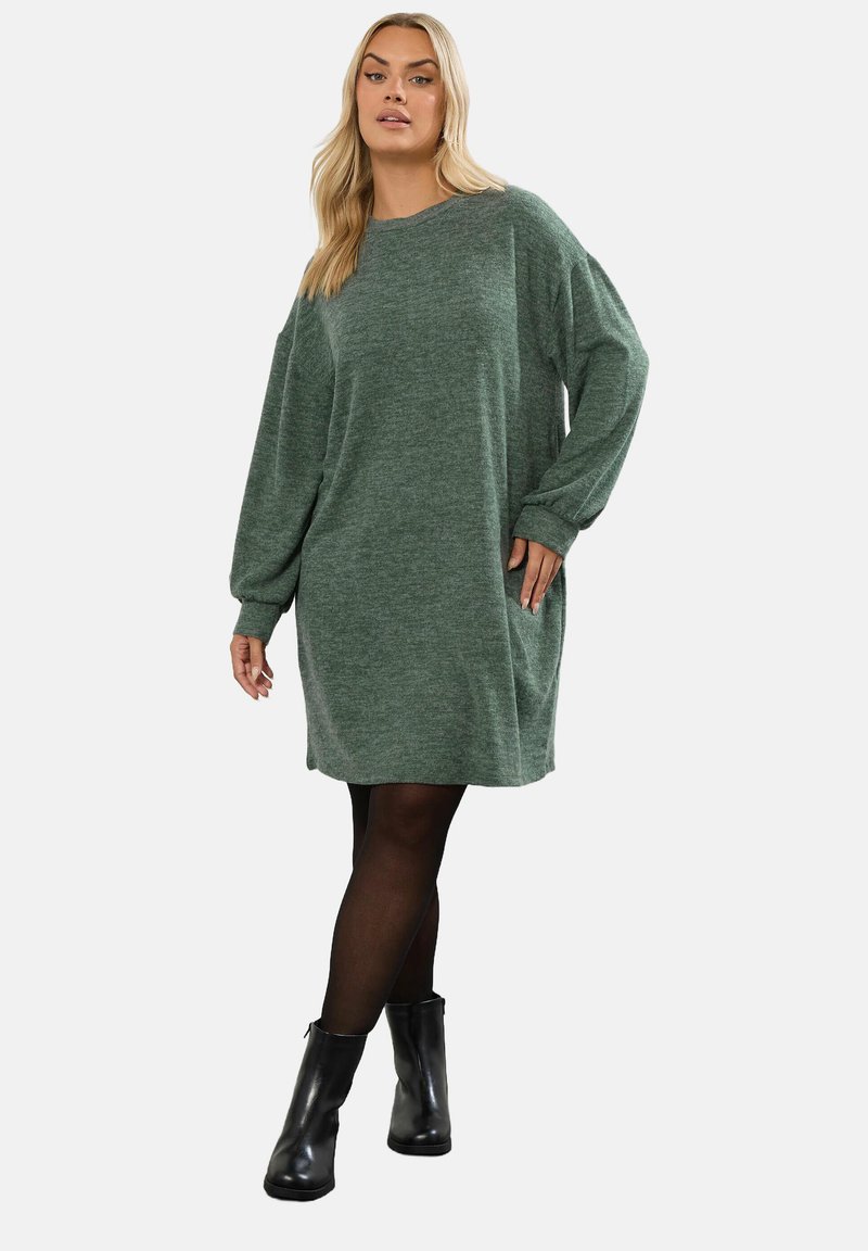 Abito maglione verde a maniche lunghe con vestibilità rilassata; presenta un collo rotondo e tasche laterali. Indossato con stivaletti neri alla caviglia.