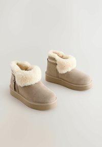 Beige Knöchelstiefel aus Wildleder mit einem flauschigen, cremefarbenen Fellkragen, strukturiertem Seitenreißverschluss und einer dicken Gummisohle.
