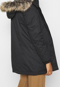 Black parka with a faux fur-lined hood, a zipper front, and ribbed cuffs. Features side pockets and a mid-length cut. Smooth texture.