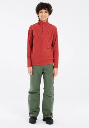 Niño sonriente con pelo rizado que lleva un forro polar rojo con cremallera hasta el pecho, pantalones verdes de exterior y zapatos negros, de pie sobre un fondo blanco.