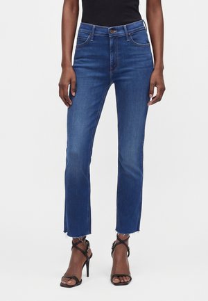 Femme portant un jean droit taille mi-haute bleu avec des ourlets effilochés et des sandales noires à talons hauts et brides, debout devant un fond uni.