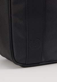 Schwarze Stofftasche mit einem strukturierten Muster, glatten schwarzen Akzenten und einem auf der Seite eingeprägten Logo. Rechteckige Form mit einem strukturierten Design.