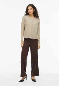 Smėlio spalvos megztinis kardiganas su priekinėmis sagomis, ribinėmis rankogaliais ir apačia, derinamas su tamsiai rudos spalvos plačiomis kelnių kojomis ir juodais aukštakulniais batais.