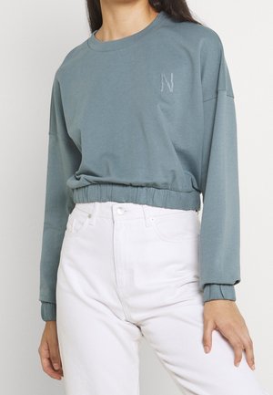 Donna che indossa una felpa grigia cropped a manica lunga con orlo elastico e pantaloni bianchi a vita alta.