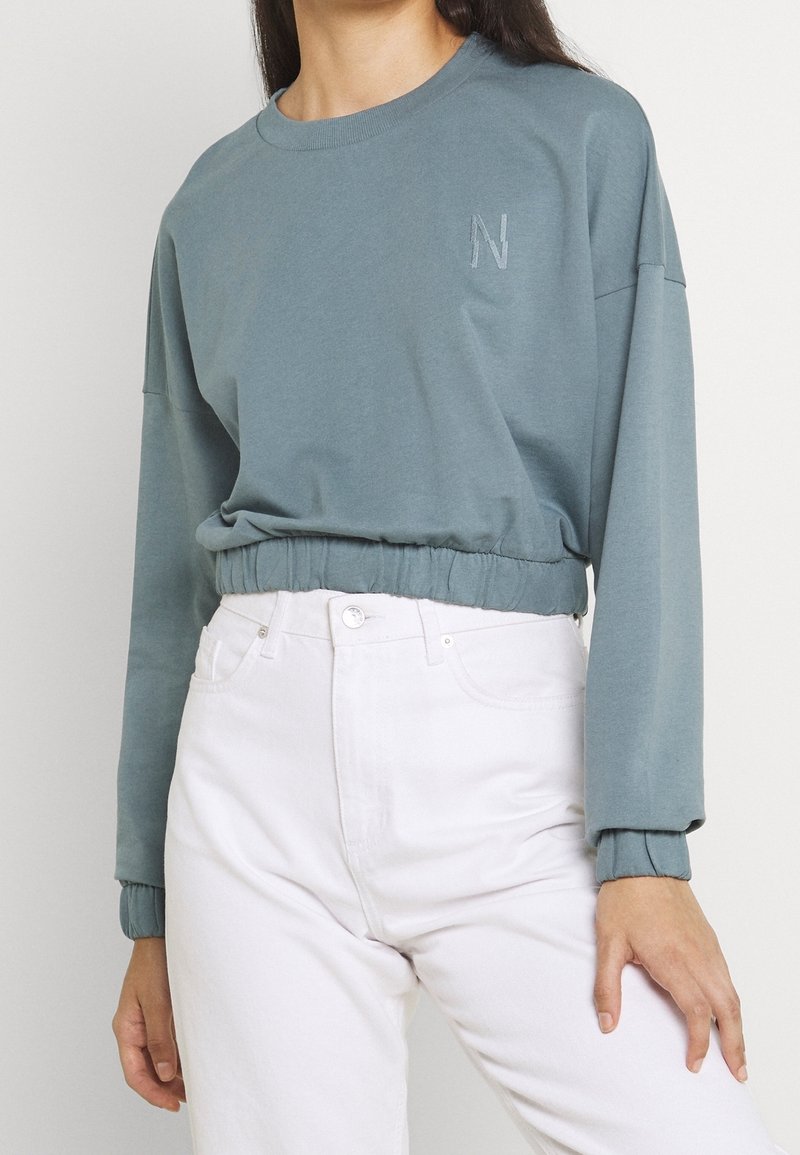 Femme portant un sweat-shirt gris court à manches longues avec un ourlet élastique et un pantalon blanc taille haute.