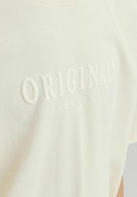 Svijetlosmeđi pamučni majica s povišenim bijelim natpisima "ORIGINALS" i "JACK & JONES", s okruglim izrezom i opuštenim krojem.