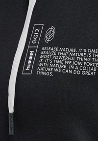 Czarna tkanina z białym napisem „hummel GG12” i wiadomością o łączeniu sił z naturą, wraz z białym sznurkiem z czarnym końcem.