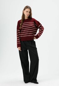 Bordeauxrode gestreepte trui met crèmekleurige horizontale lijnen, voorzien van een losse pasvorm en geribbelde zoom. Gecombineerd met losse zwarte broek en zwarte schoenen.