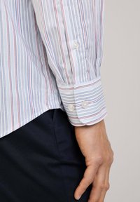 Katoenen shirt met verticale blauwe, rode en witte strepen; voorzien van een geknoopte manchet met twee knopen en een rechte zoom.