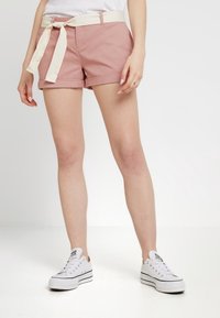 Shorts en coton rose avec un ourlet replié et une ceinture en coton crème. Portés avec des baskets blanches, avec des accents noirs. Design simple, sans motifs.