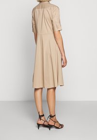 Beige knälång klänning med korta ärmar, knappade manschetter och en utsvängd kjol. Bärs med svarta sandaletter med remmar och en slät textur.