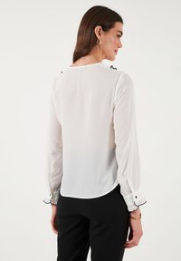 White, sheer long-sleeve blouse with black trim at cuffs and collar. Features a rounded hem and button detailing at shoulders.