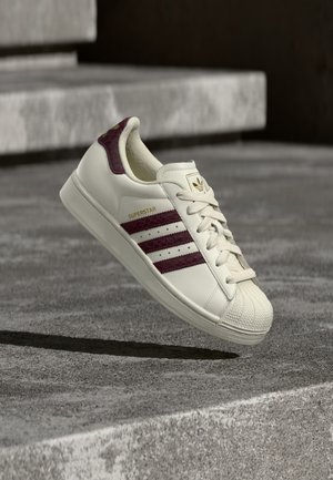 Baskets en cuir blanc avec accents bordeaux, présentant le design classique à trois bandes, embout en caoutchouc et marquage doré "SUPERSTAR".