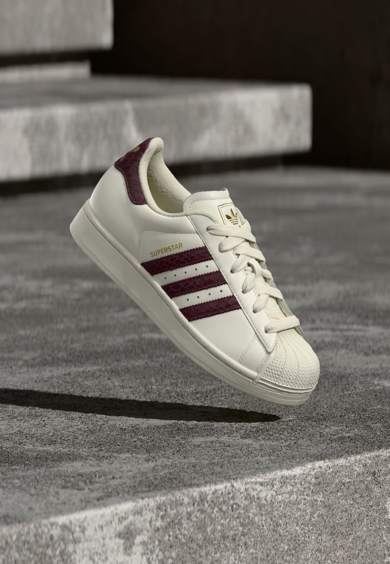 Baskets en cuir blanc avec accents bordeaux, présentant le design classique à trois bandes, embout en caoutchouc et marquage doré "SUPERSTAR".