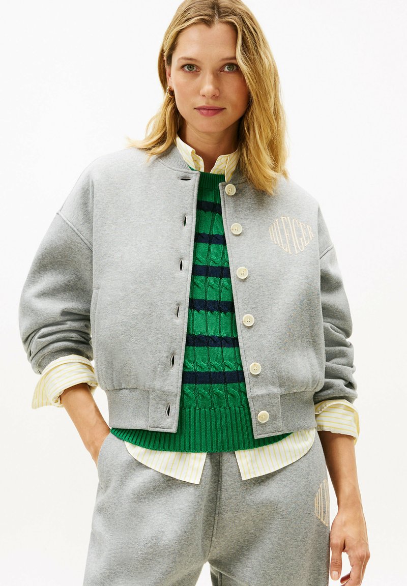 Mujer con chaqueta gris abotonada y pantalones, suéter de rayas verde y azul marino y camisa de rayas amarillo claro, de pie con las manos en los bolsillos.