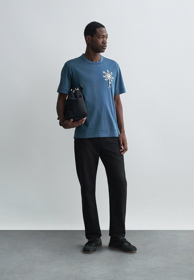 Camiseta de algodão azul com um design floral branco no lado esquerdo do peito, combinada com calças pretas e sapatos pretos, segurando uma bolsa preta.