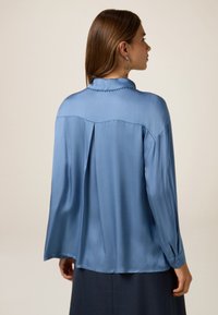 Blusa de satén azul claro con cuello, mangas largas y un pliegue en la parte posterior. Cuenta con puños abotonados y una textura suave y brillante.