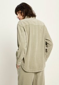 Person with shoulder-length brown hair wearing a loose beige corduroy shirt and matching pants, hands in pockets, looking to the side.