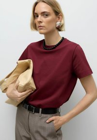 Bordeaux katoenen t-shirt met korte mouwen, gecombineerd met een beige tote bag en grijze geblokte broek. Zwarte kralenketting en zilveren oorbellen.