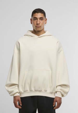 Sweat à capuche surdimensionné couleur crème en tissu doux, avec une poche avant et une capuche à cordon. Logo discret blanc sur la poitrine.