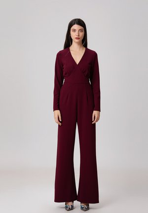Combinaison bourgogne à manches longues, décolleté en V profond, taille plissée et coupe à jambes larges. Associée à des chaussures à talons hauts à motifs.