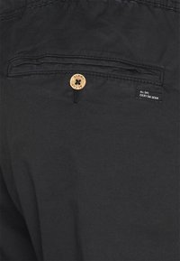 Zwarte spijkershorts met een achterzak voorzien van een bruine knoop en een label met de tekst "ALL DAY EVERY DAY DENIM." Soepele textuur en casual ontwerp.