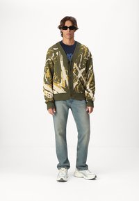 Strikket cardigan i oliven med gult og hvitt abstrakt mønster, med v-hals, knapper og ribbestrikkede mansjetter, kombinert med lyseblå jeans.