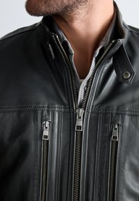 Homme portant une chemise grise superposée et une veste en cuir noire avec trois fermetures éclair verticales visibles et un col à pression.