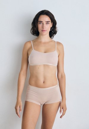 Woman with short dark hair wearing nude bra and boyshort underwear standing against plain light background facing forward.