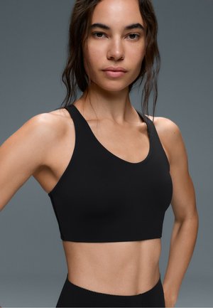 Jeune femme aux longs cheveux foncés porte un soutien-gorge de sport noir, debout les mains sur les hanches devant un fond gris.
