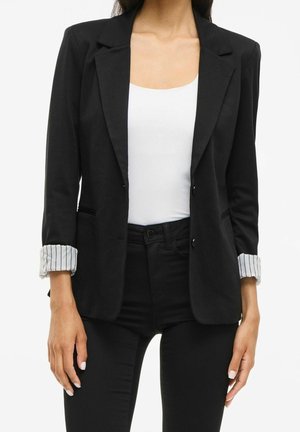 Kvinde iført en sort blazer med stribet manchetforing over en hvid top og sorte bukser, stående mod en ensfarvet hvid baggrund.