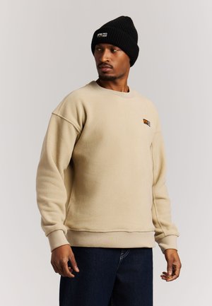 Man draagt een beige fleece sweater en een zwarte gebreide muts, kijkt opzij tegen een effen achtergrond.