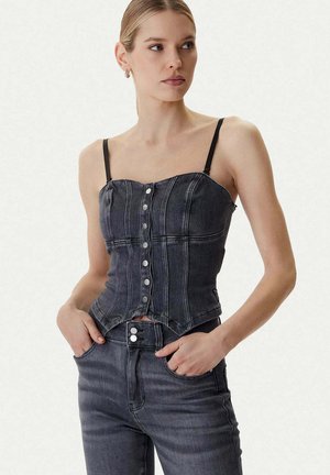Donna che indossa un top corsetto di jeans grigio scuro aderente con bottoni e spalline sottili, abbinato a jeans a vita alta, con le mani nelle tasche, che guarda di lato.
