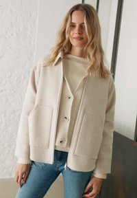 Cream-colored wool blend jacket with a collared neckline, two front pockets, and a zip closure, layered over a neutral knit sweater.