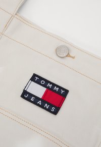Beige tyg med synlig sömnad; har en navy, vit och röd broderad logotyp som säger "TOMMY JEANS" ovanför en metallknapp.