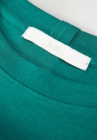 Nahaufnahme eines türkisfarbenen T-Shirts mit einem weißen eingenähten Etikett, auf dem der verblasste Text "N.82" steht.