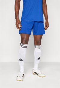 Άνδρας αθλητής που φορά μπλε αθλητικό σορτς, λευκές ποδοσφαιρικές κάλτσες με επιγονατίδες και λευκά παπούτσια με μαύρες ρίγες, στέκεται σε απλό φόντο.