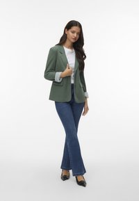 Blazer verde con rever a punta, polsini a quattrofoglie, abbinato a una camicia bianca, jeans a zampa blu e décolleté nere a punta.