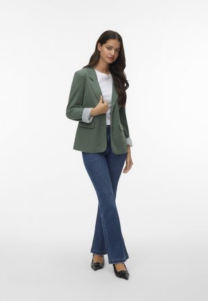 Grön kavaj med notched lapels, kvadratisk textur på manschetterna, i kombination med en vit skjorta, blå utställda jeans och svarta spetsiga klackar.