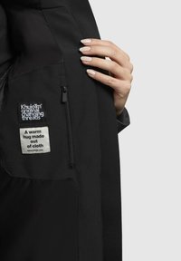 Intérieur de veste noire avec une poche zippée, comportant deux étiquettes : une de marque et une indiquant "Une étreinte chaleureuse faite de tissu." Texture lisse.