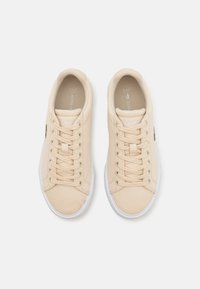 Beige kanvas sneakers med hvite gummisåler, flate lisser, og en liten grønn krokodillelogo på siden. Jevn tekstur, rund tådesign.