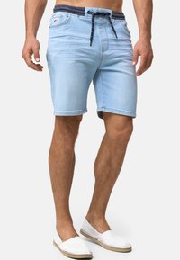 Lyseblå denimshorts med snor i livet i marineblå, med fem lommer og rett snitt. Brukt med hvite slip-on sko.