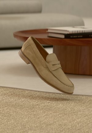 Beiger Wildleder-Slipper mit Pennyriemen-Detail, schwebend über einem strukturierten beigen Teppich, im Hintergrund ein runder Holzcouchtisch und gestapelte Bücher.
