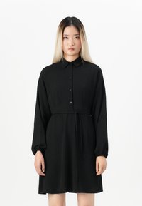 Young woman with straight blond-black hair wearing a long-sleeve black button-up dress with a tied waist, standing against a white background.