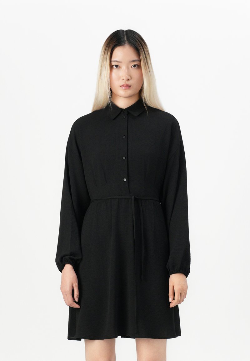 Young woman with straight blond-black hair wearing a long-sleeve black button-up dress with a tied waist, standing against a white background.