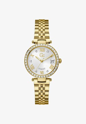 Montre-bracelet en métal doré avec un cadran rond, un fond blanc, des chiffres romains et une lunette ornée de cristaux. Elle comporte un affichage de la date et un bracelet.