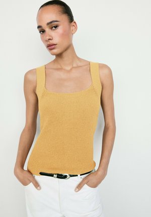 Donna con capelli tirati indietro che indossa un top giallo senape di maglia infilato in pantaloni bianchi con cintura nera, in piedi davanti a uno sfondo semplice.